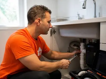 Sanitario con remera naranja arreglando con una llave la cañería debajo de la pileta de una cocina