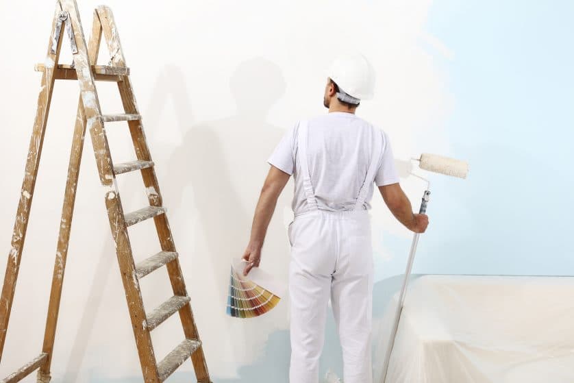Pintor con un rodillo y una carta de colores frente a una pared, con los muebles cubiertos con nylon